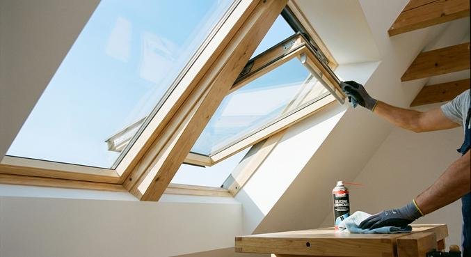 Close-up of hands cleaning and maintaining a modern wooden roof window in a bright attic.