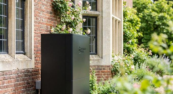 Modern air-source heat pump installed outside a traditional brick house with green plants in a garden.